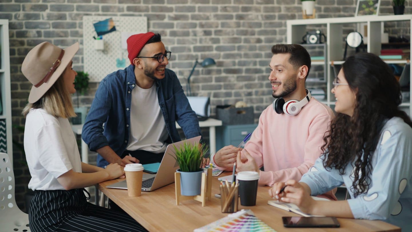 Cuatro profesionales diversos colaborando alegremente alrededor de una mesa de oficina.